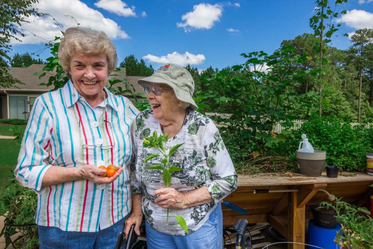 Two smiling women gardening, surrounded by plants and vegetables.
